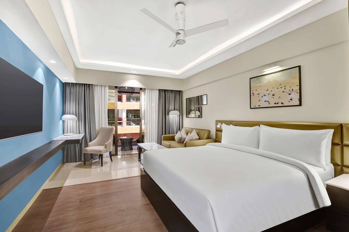 A hotel room with a large bed, a desk, a chair, a TV, and a ceiling fan.