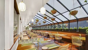 Un ampli restaurant amb sostres alts, plantes penjants i una llarga zona de menjador amb cadires de vimbles i sotsplats ratllats.