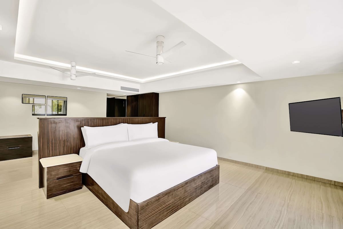 A modern bedroom with a large bed, a wooden nightstand, a ceiling fan, and a flat-screen TV mounted on the wall.