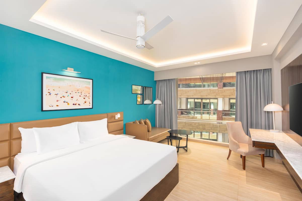 A modern hotel room with a large bed, a desk, a chair, a ceiling fan, and a view of the city through large windows.