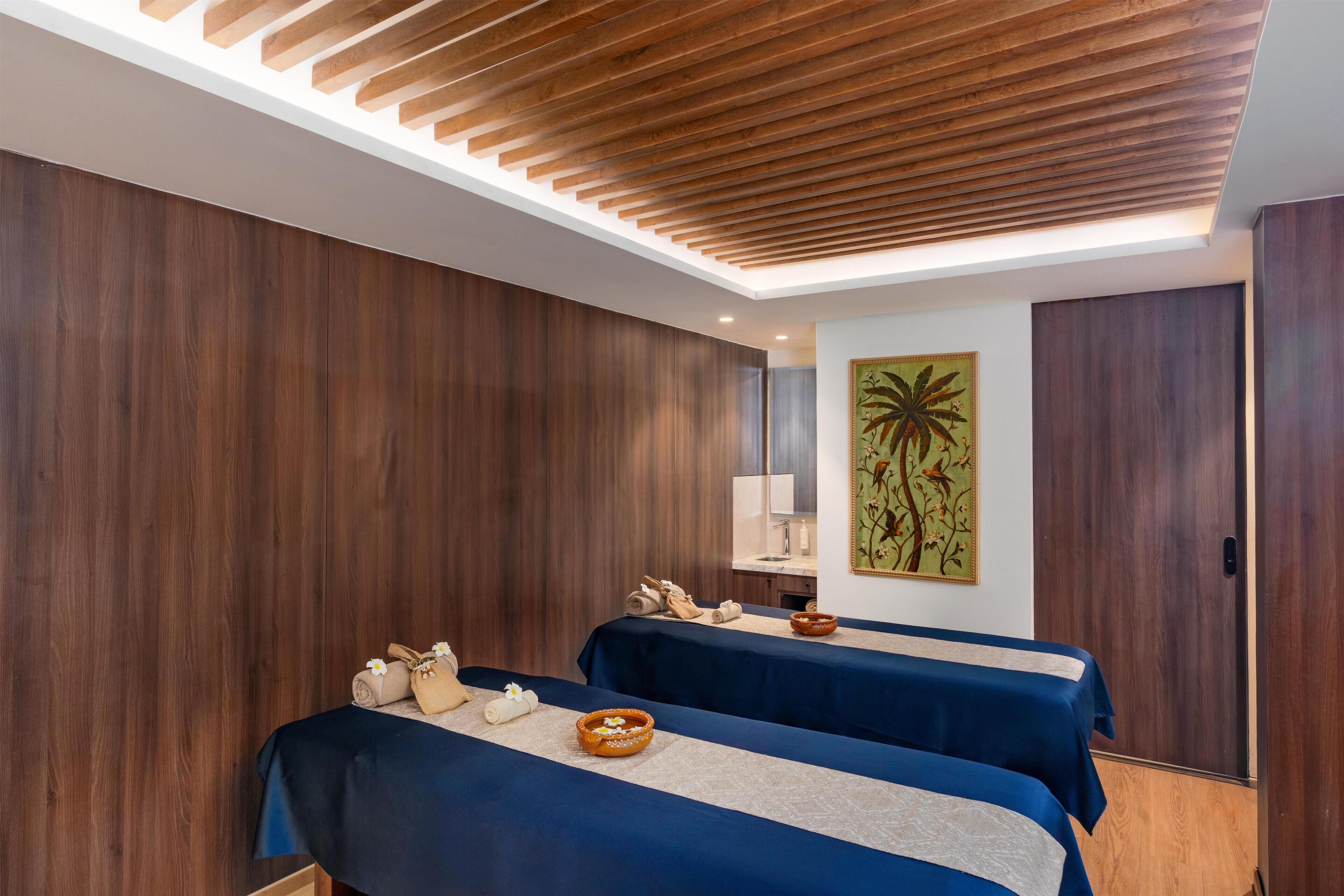 A spa room with two massage tables, wooden panel walls, a ceiling with wooden slats, and a painting of a tree.