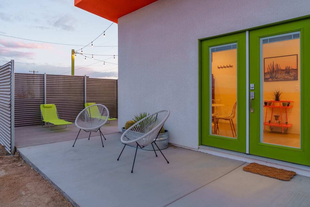 En moderne udendørs terrasse med to hvide wirestole, en grøn dør og et lille indendørs område med en plante og en reol.