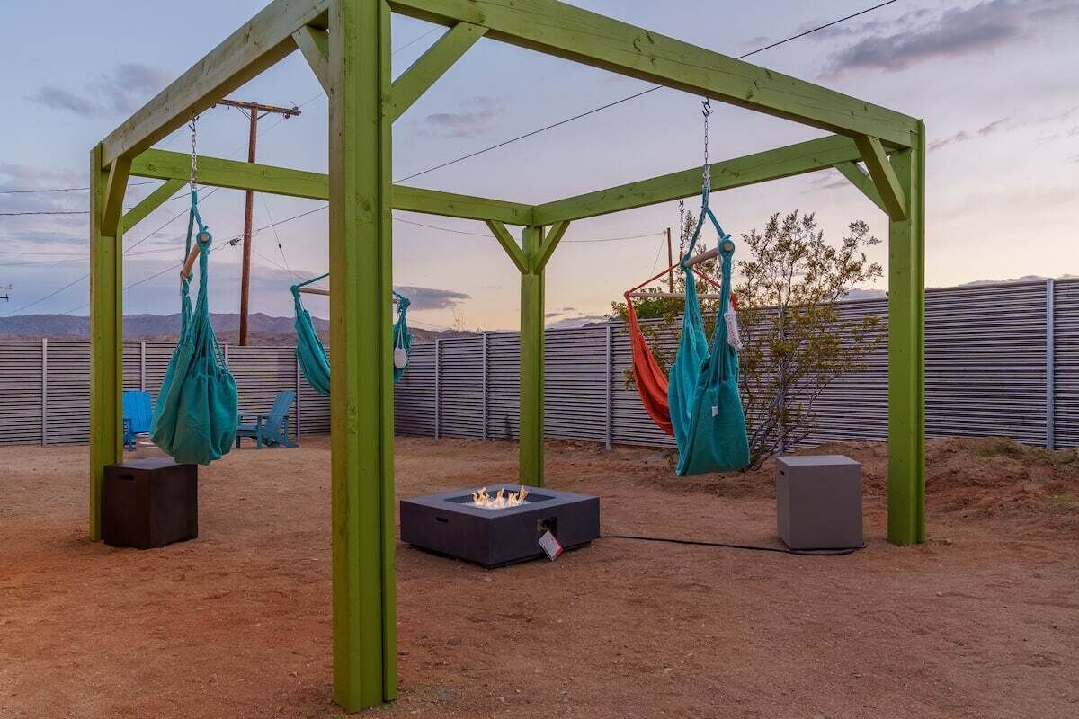 A wooden pergola structure with a fire pit in the center, surrounded by green and blue hammocks.
