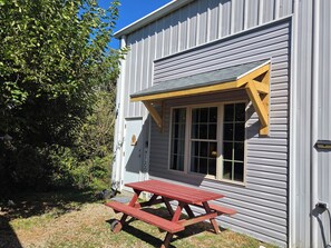 A building with a wooden picnic table outside, a window with a triangular awning, and a door with a 'WELCOME' sign.