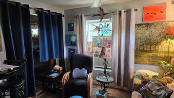 Un salon chaleureux avec un fauteuil noir, une petite table ronde et un canapé. On y trouve un ventilateur de plafond, une lampe et une fenêtre avec des rideaux.