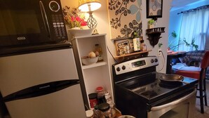 Eine kompakte Küche mit Kühlschrank, Mikrowelle, Herd und Backofen. Ein Holzschemel mit einem Topf darauf und verschiedene Küchenutensilien auf der Arbeitsfläche.