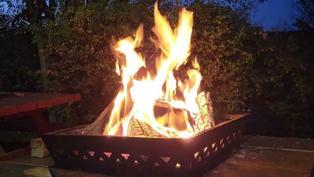 Ein Lagerfeuer mit Flammen, umgeben von einer Terrasse mit Tisch und Bänken, vor einer Kulisse aus Bäumen und blauem Himmel.