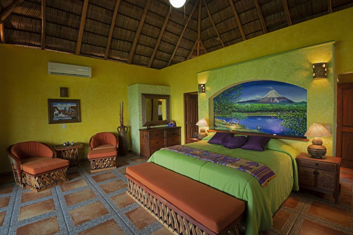 A room with a bed, two chairs, a bench, a nightstand, a lamp, a mirror, and a painting of a landscape.