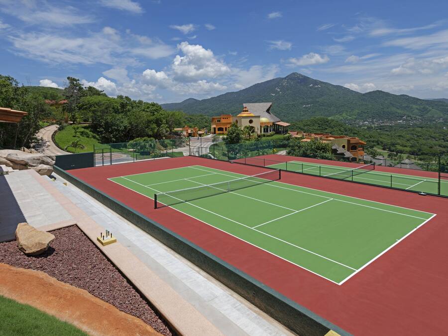 Una cancha de tenis con una red, rodeada de una superficie roja y verde, con montañas y un cielo despejado al fondo.