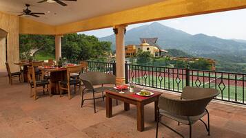 Una terraza con mesas y sillas, una cancha de tenis al fondo y vistas a la montaña.