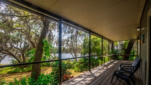 Une véranda couverte avec un plancher en bois, deux chaises noires et une vue sur un lac entouré d’arbres.