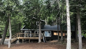 A cabin with a deck, surrounded by trees. - A Hidden Gem with Sandy Shore Memories and Island Views. (Township of Muskoka Lakes)
