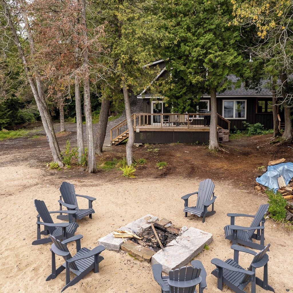 A backyard with a fire pit surrounded by Adirondack chairs, a wooden deck, and a house with a staircase leading to a deck.