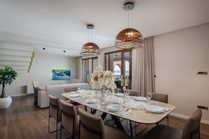 Un espace de salle à manger moderne, avec une longue table dressée pour six personnes, entourée de chaises. Deux lampes suspendues sont placées au-dessus de la table, et une grande fenêtre avec des rideaux se trouve à droite.