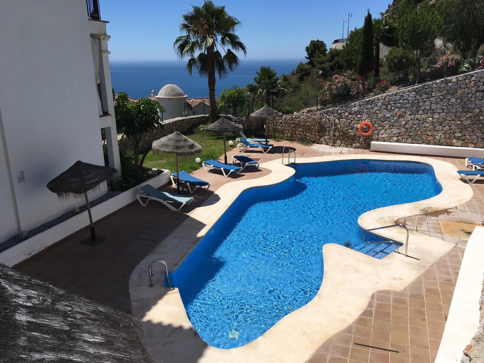 A swimming pool with a curved edge, surrounded by lounge chairs and umbrellas, with a view of the sea and a stone wall in the background.