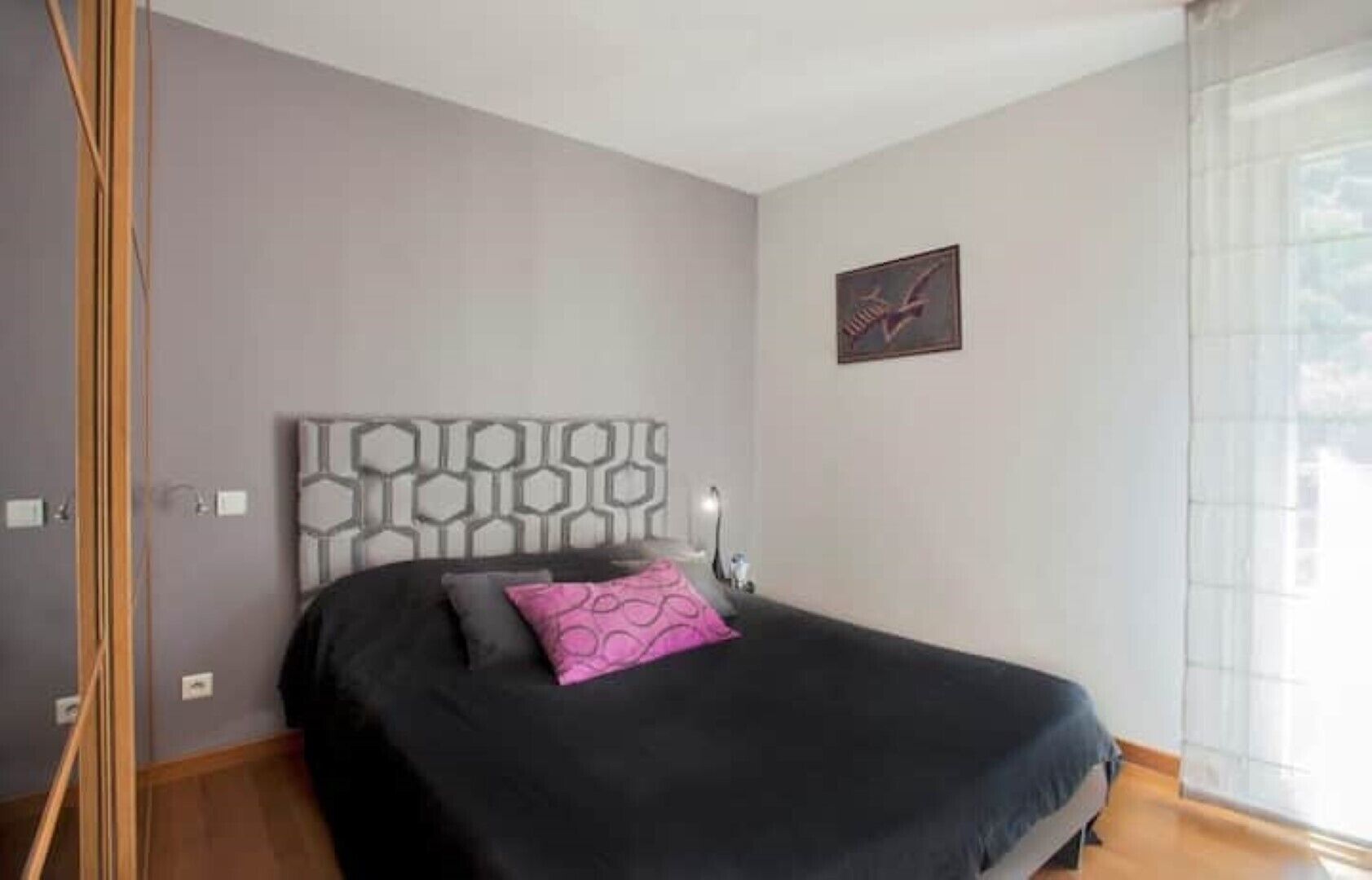 A bedroom with a bed, a headboard with a geometric pattern, a bedside table, and a window with curtains.