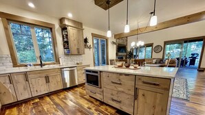 Een moderne keuken met houten kastjes, een vaatwasser, een oven en een groot eiland met een spoelbak. Boven het eiland hangen hanglampen en er is een accentmuur van steen.
