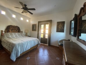 A bedroom with a bed, a ceiling fan, a door, and a small table with a lamp.