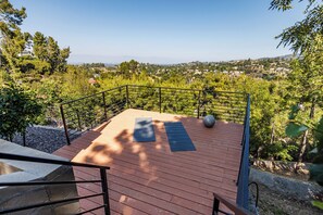 Una terraza de madera con un colchoneta de ejercicio negro y un objeto esférico grande, rodeada por una barandilla metálica, con vista a un paisaje verde.