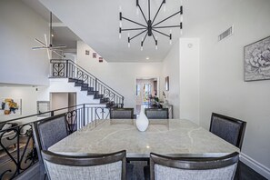 A dining area with a marble table, upholstered chairs, a modern chandelier, and a staircase leading to an upper level.