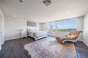 Un dormitorio con una cama grande, una zona de estar con una silla, una mesita de noche y vistas al exterior a través de un ventanal.
