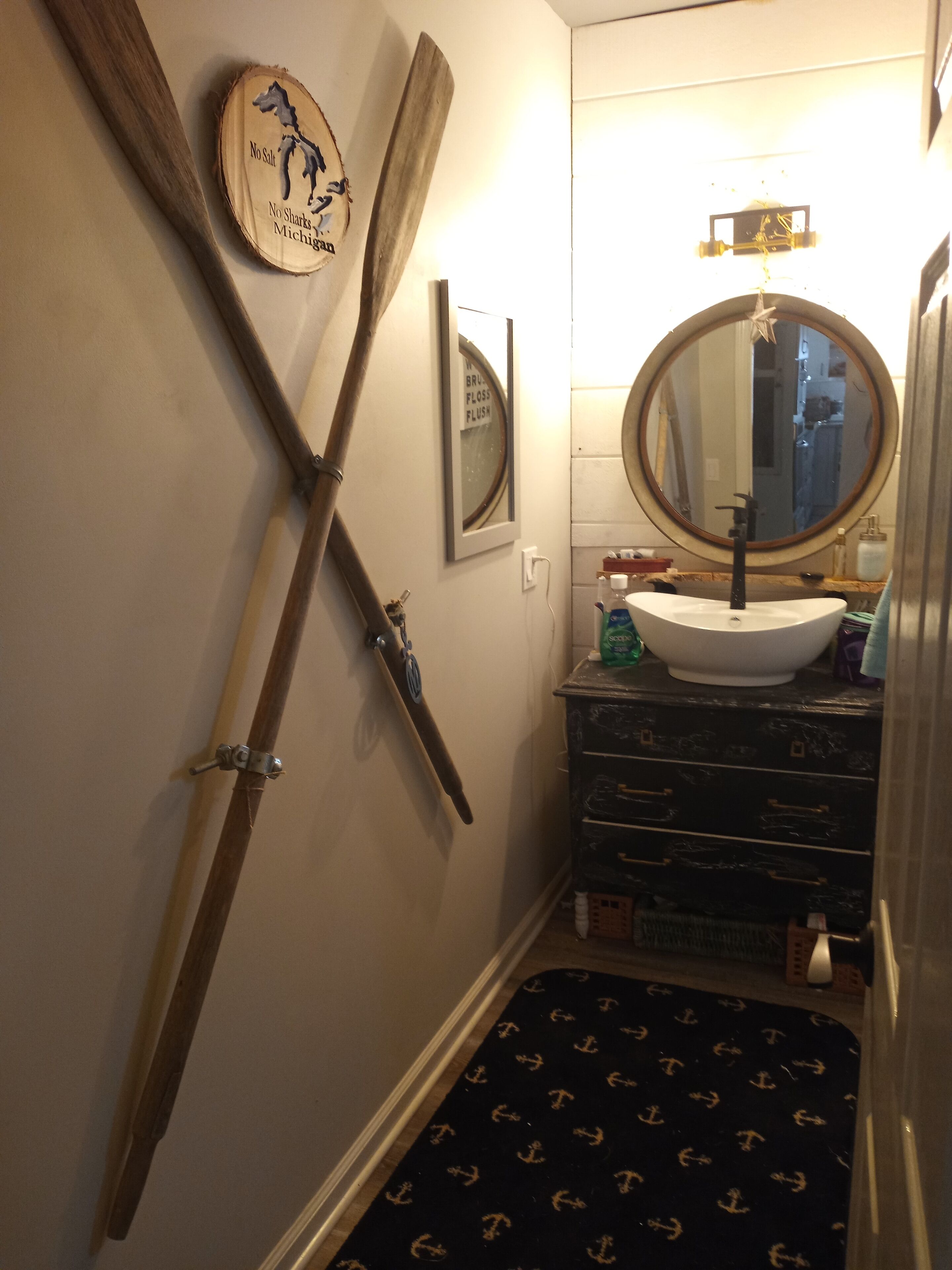 Une salle de bain avec une pagaie en bois appuyée contre le mur, un miroir rond au-dessus d’un lavabo et un petit tapis orné de motifs d’ancre.