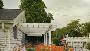 A house with a porch, surrounded by a garden with orange flowers and green plants. - UPPER 3 rm.  studio B&B steps from beach & farm markt/shop'g.; dogs ok w/fee.  (Port Austin)