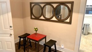 Een kamer met een houten vloer, een rode tafel met twee zwarte stoelen, een spiegel aan de muur en een deur.