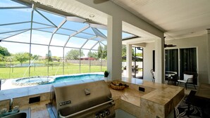 An outdoor kitchen with a grill, a pool, and a view of a grassy yard. - Luxury 4 BR/3BA Waterview Home w/ Pool + Spa with work space (Bradenton)