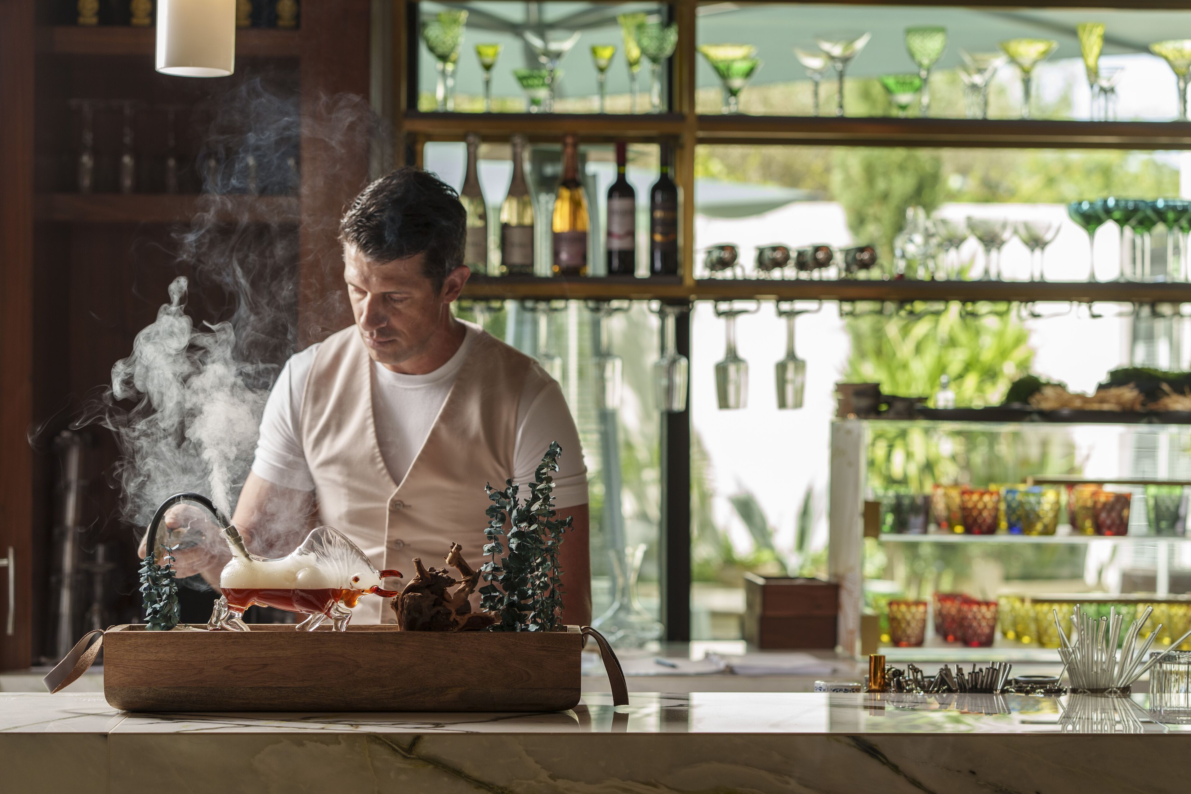 A person working with a smoking device at a bar counter, surrounded by shelves of glassware and bottles.