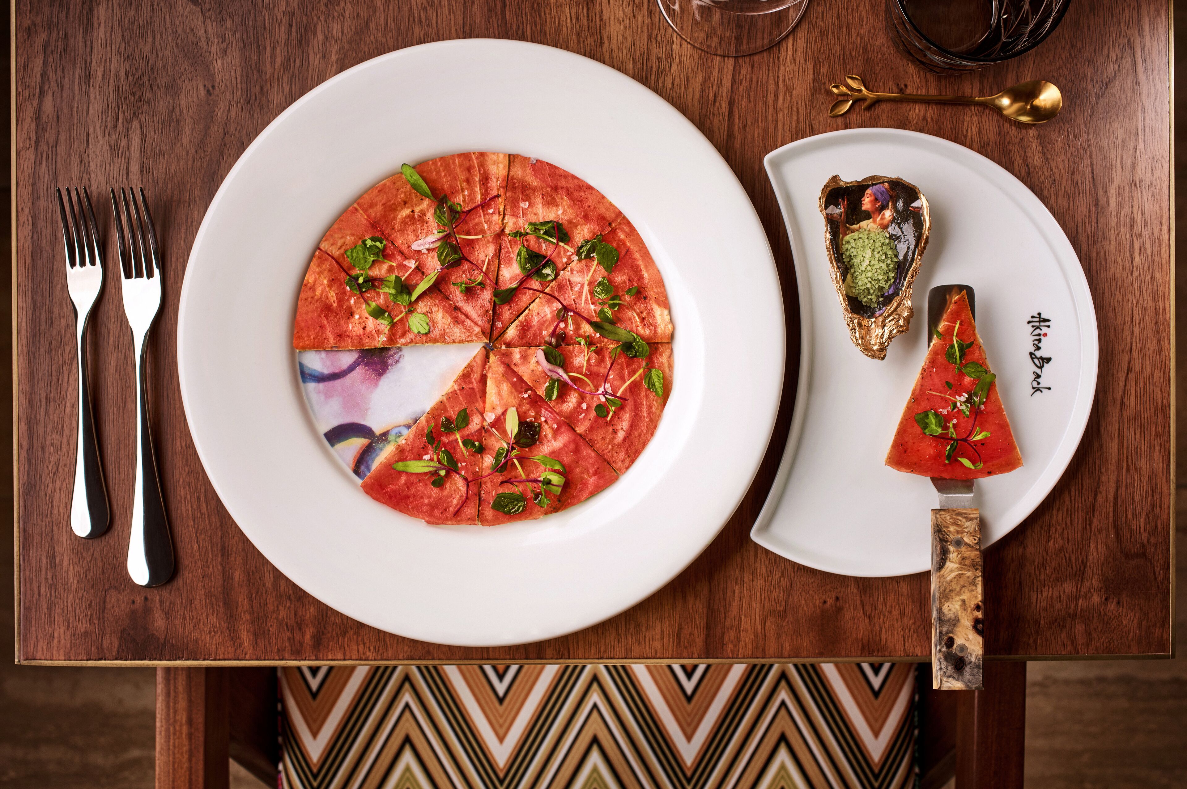A dining table with a pizza, a carved watermelon, and a plate with a name.