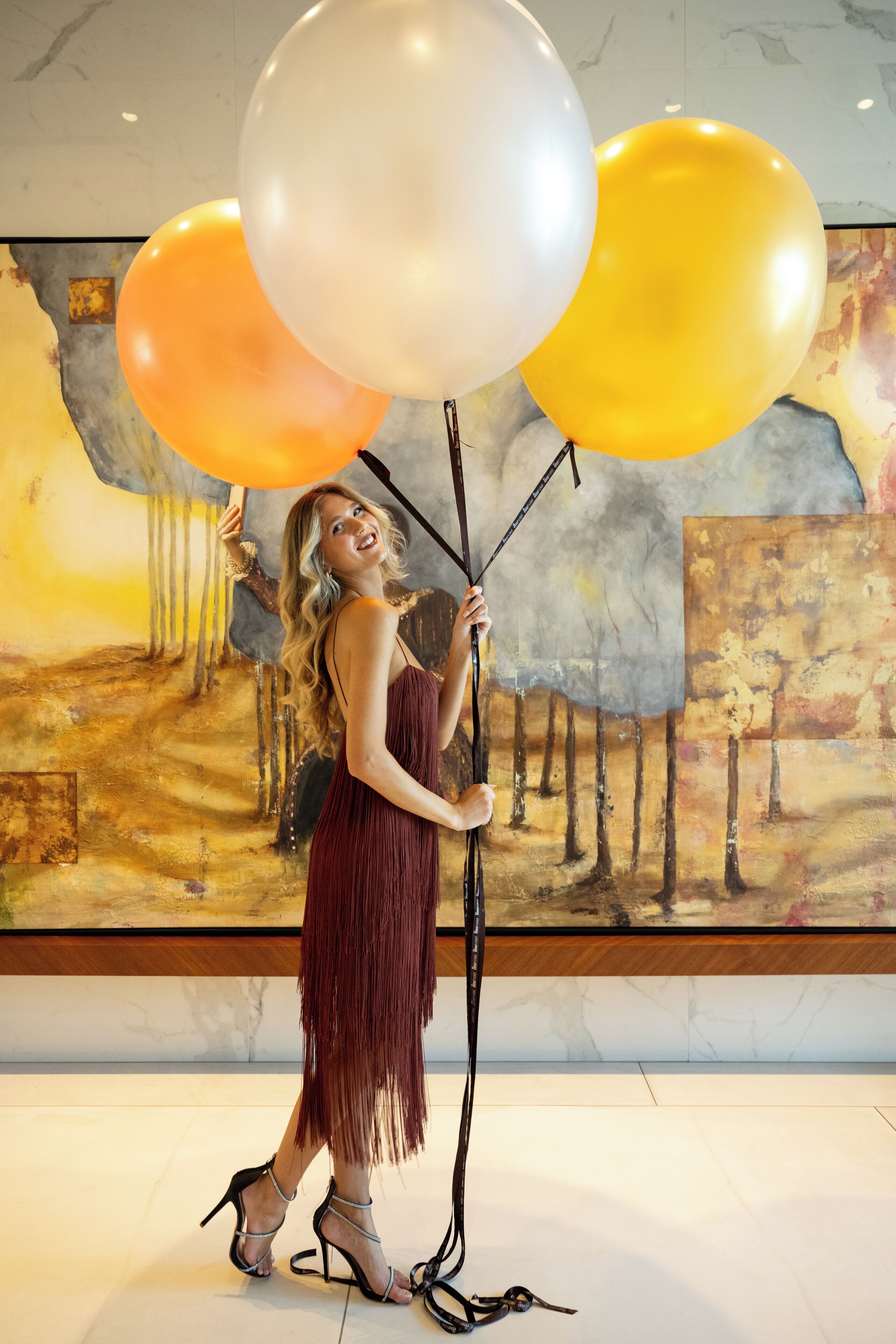 Una persona sosteniendo globos en una habitación con piso de mármol y un cuadro abstracto grande en la pared.