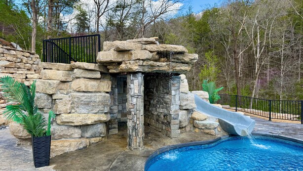 A swimming pool with a stone waterfall and a slide, surrounded by a stone wall and a metal fence. - Private Retreat For You w/HEATED Pool, Private Dock,  Activity gym, 100 acres! (Galena)