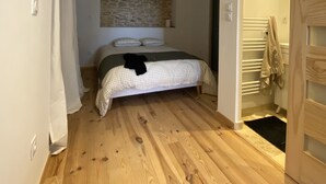 Une chambre avec un plancher en bois, un lit recouvert d’une couette à carreaux, un mur en pierre et une porte en bois.