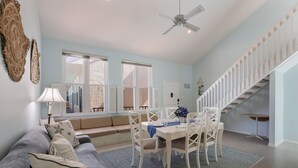 A spacious living room with a high ceiling, a dining area with white chairs, a blue accent wall, and a staircase with white railings.