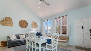 A dining area with a white table, four chairs, a blue tablecloth, and a vase with flowers. There is a grey sofa with cushions and a woven basket on the floor.