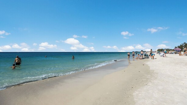 A beach with clear blue water, people swimming and walking along the shore, and beachgoers relaxing on the sand.