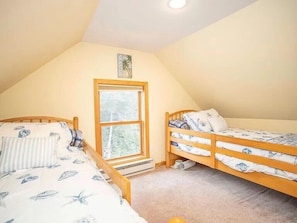 A small, cozy attic bedroom with two wooden bunk beds, a window with a view of trees, and a ceiling light. - Perfect Shoreline Stay – Walk to Beach, Dine, Shop & Play! (Westbrook)