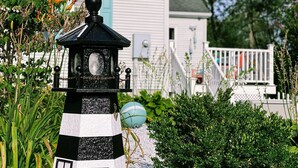 Un phare aux rayures noires et blanches, surmonté d’une petite cloche, entouré d’une végétation luxuriante et, au fond, une maison blanche avec une porte bleue.