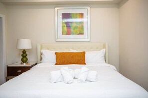 Un lit bien fait, avec des draps blancs et des serviettes pliées, un seul coussin orange et, au-dessus, un tableau abstrait encadré.