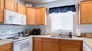 Une cuisine avec des meubles en bois, un four blanc, un four à micro-ondes, un évier et une fenêtre avec des stores.