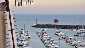 Uma marina com vários barcos atracados, um farol ao fundo e um quebra-mar rochoso.