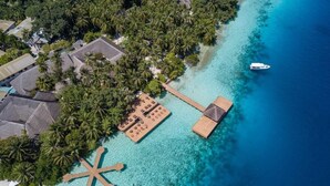Vista aérea de un resort tropical con mar turquesa cristalino, palmeras y una terraza de madera con sillones de descanso.