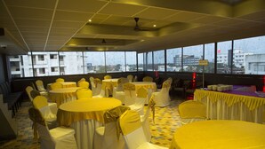 Un salón de banquetes con mesas y sillas redondas, ventanas amplias con vista a la ciudad y un ventilador de techo.