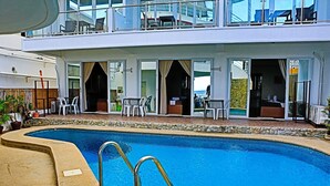 A pool area with a clear view of the pool, lounge chairs, and a building with multiple balconies. - Arkipelago Beach Resort (Puerto Galera)