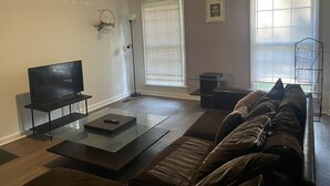 Un soggiorno con una TV a schermo piatto su un supporto nero, un tavolino da caffè in vetro, un divano in pelle marrone e una lampada da terra.
