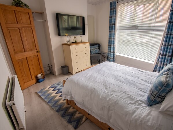 Ein Schlafzimmer mit Bett, Kommode, Fernseher, Heizkörper und einem Fenster mit Vorhängen.