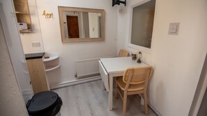 A small, clean room with a white table and two wicker chairs, a mirror, a radiator, and a black trash bin.