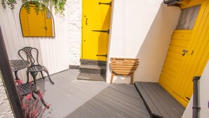 A small outdoor area with a yellow door, a wooden bench, and two metal chairs.