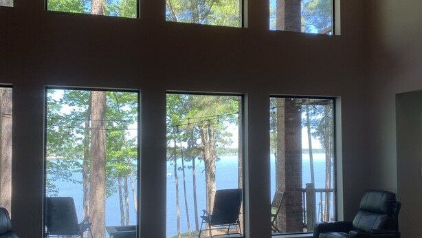 Ein geräumiges Zimmer mit großen Fenstern mit Blick auf ein Gewässer und Bäume. Zwei schwarze Ledersessel und ein weißes Sofa sind vorhanden.